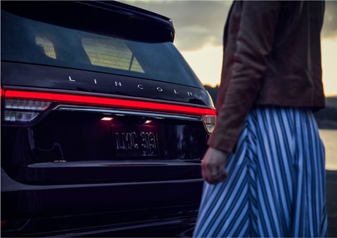 A person is shown near the rear of a 2023 Lincoln Aviator® SUV as the Lincoln Embrace illuminates the rear lights | Oliver Lincoln in Plymouth IN