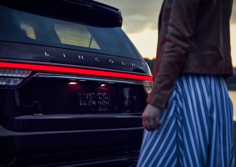 A person is shown near the rear of a 2024 Lincoln Aviator® SUV as the Lincoln Embrace illuminates the rear lights | Oliver Lincoln in Plymouth IN